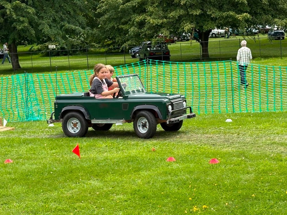 Two children sharing a mini Land Rover on the roped-off course at a classic car show