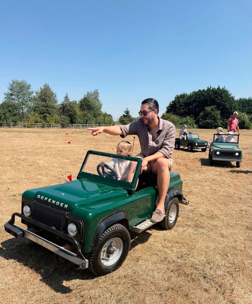 A grown-up riding alongside a small child at the wheel of a mini Land Rover, with the rest of the fleet out on the course behind