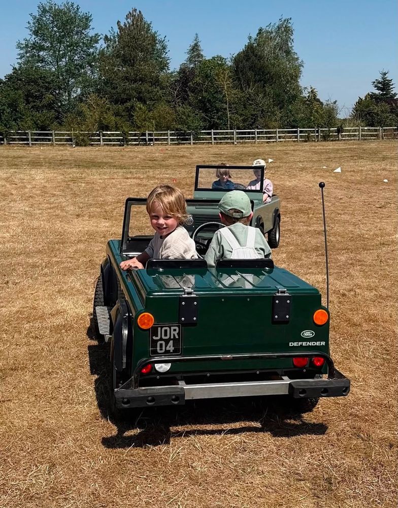 Two grinning children in JOR 04 following another mini Land Rover across a sunlit field
