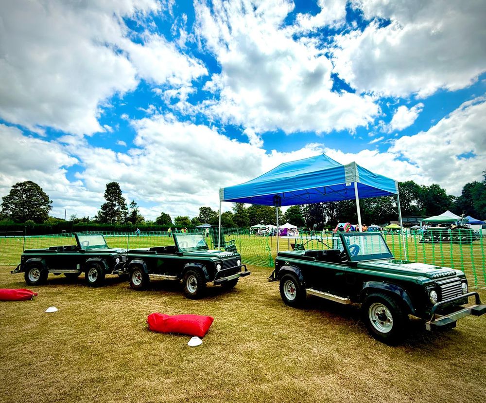 Three mini Land Rovers lined up beside the gazebo at a country show, under a big cloudy sky