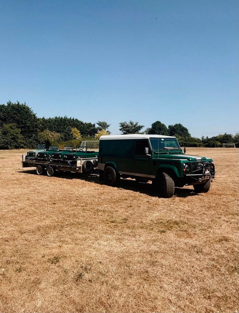 A full-size Land Rover Defender towing a trailer loaded with three mini Land Rovers across a field